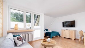 A modern, well-lit room with a large window, a bed with a teal bedspread, a wooden cabinet, and a television on the wall.