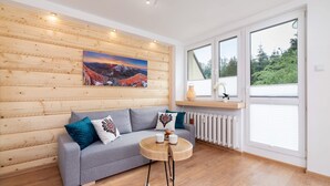 A modern living room with a grey sofa, wooden panel walls, a round wooden coffee table, and a large window with blinds.