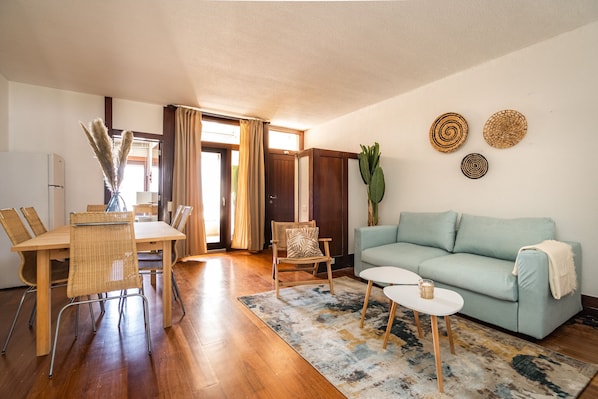 A living room with a light blue sofa, a dining table with chairs, a coffee table, and a large plant.