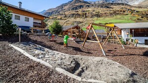 Un parque infantil con estructuras de madera, un edificio de techo inclinado y montañas al fondo.