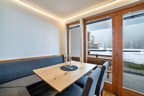 Een modern interieur met een houten tafel en een bank, grote ramen met uitzicht op een besneeid landschap.