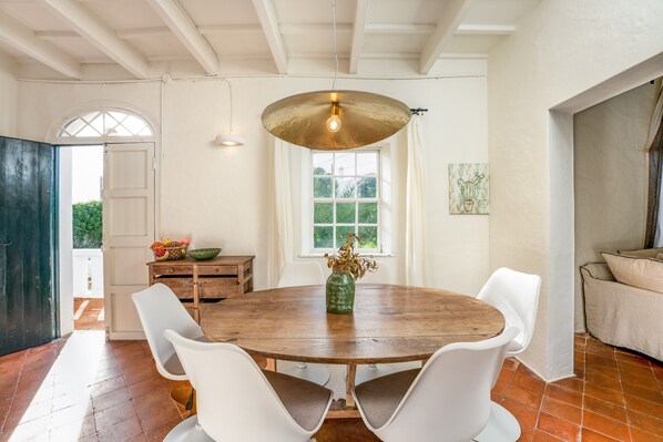 A dining area with a wooden table, white chairs, a hanging light fixture, a window with a view, and a door leading outside.