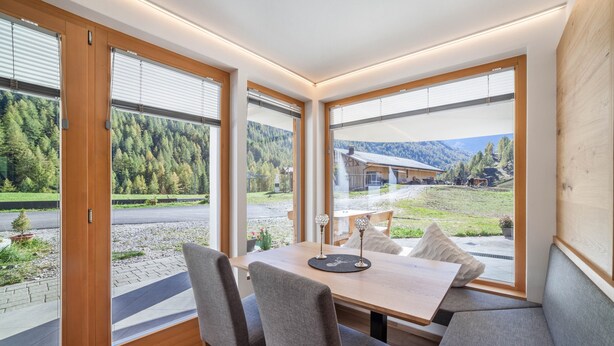 A modern dining area with a table and chairs, large windows with blinds, and a view of a forested mountain area. - Apartment 'Lazins' with Private Terrace & Wi-Fi (Pfelders)