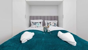 Un lit bien fait, recouvert d’une courtepointe turquoise, avec des oreillers blancs et une bouteille de champagne posée au centre.
