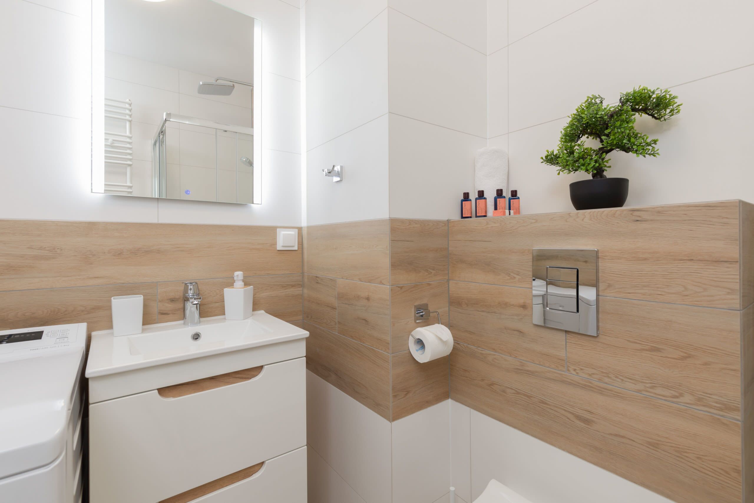 Un baño moderno con tocador de madera, espejo, lavamanos, dispensador de papel higiénico y una planta en maceta.