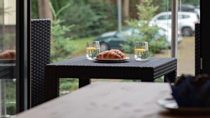 Une table à manger avec un croissant et deux verres d’eau, une chaise en osier et, par la fenêtre, une vue sur la verdure et les voitures garées.