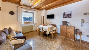 A cozy room with a wooden ceiling, a dining table with chairs, a sofa, a TV mounted on the wall, and a window with a view of mountains. - Eichernhof Apartment Kirschblüte – Delicious Vital Breakfast and Dinner Available on Request (Verano)