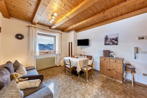 Una habitación acogedora con techo de madera, una mesa de comedor con sillas, un sofá, un televisor fijado a la pared y una ventana con vistas a montañas.