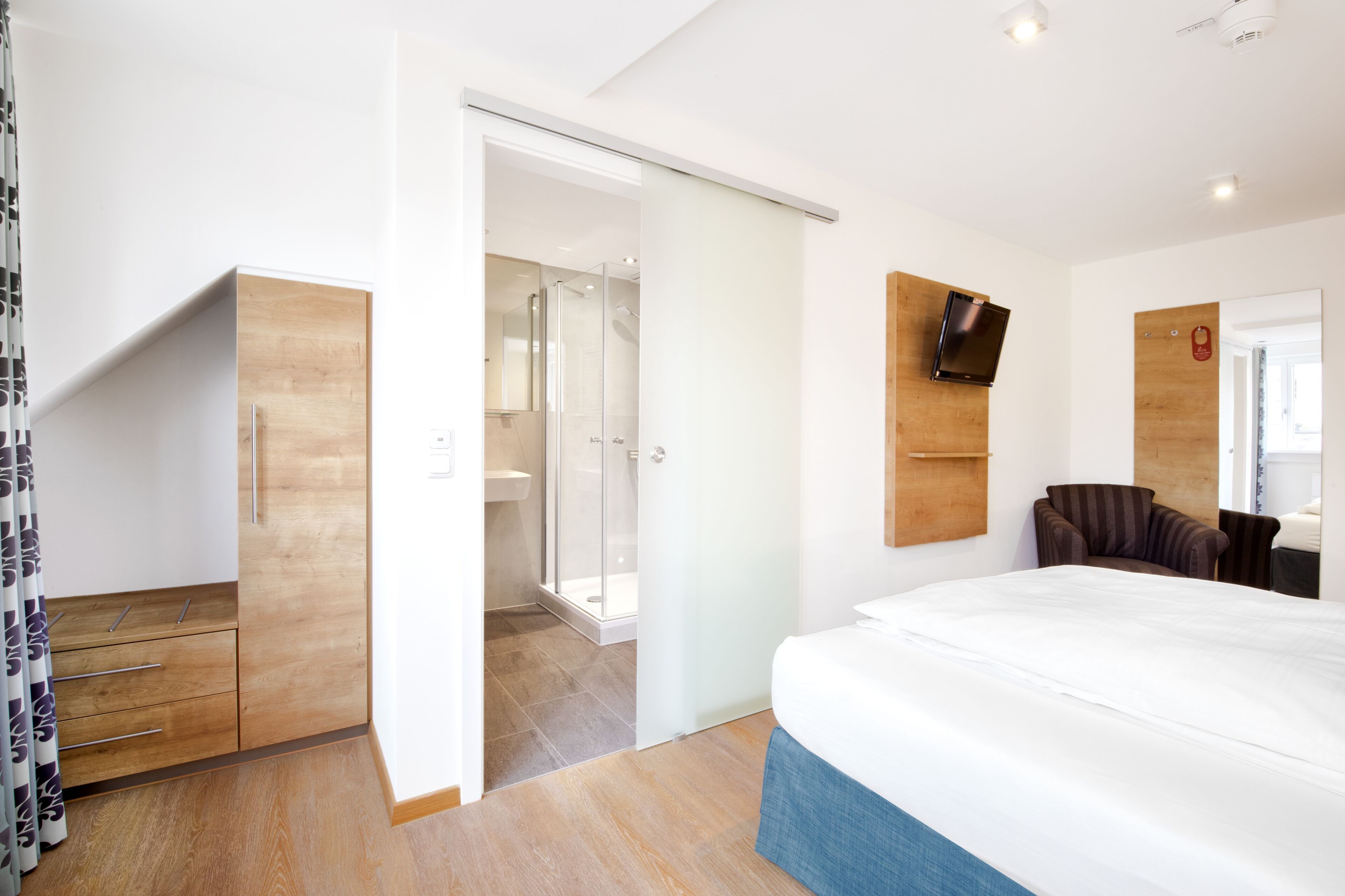 Ein modernes Schlafzimmer mit einem großen Bett, einem Holzschrank, einer Duschkabine und einem an der Wand befestigten Fernseher.