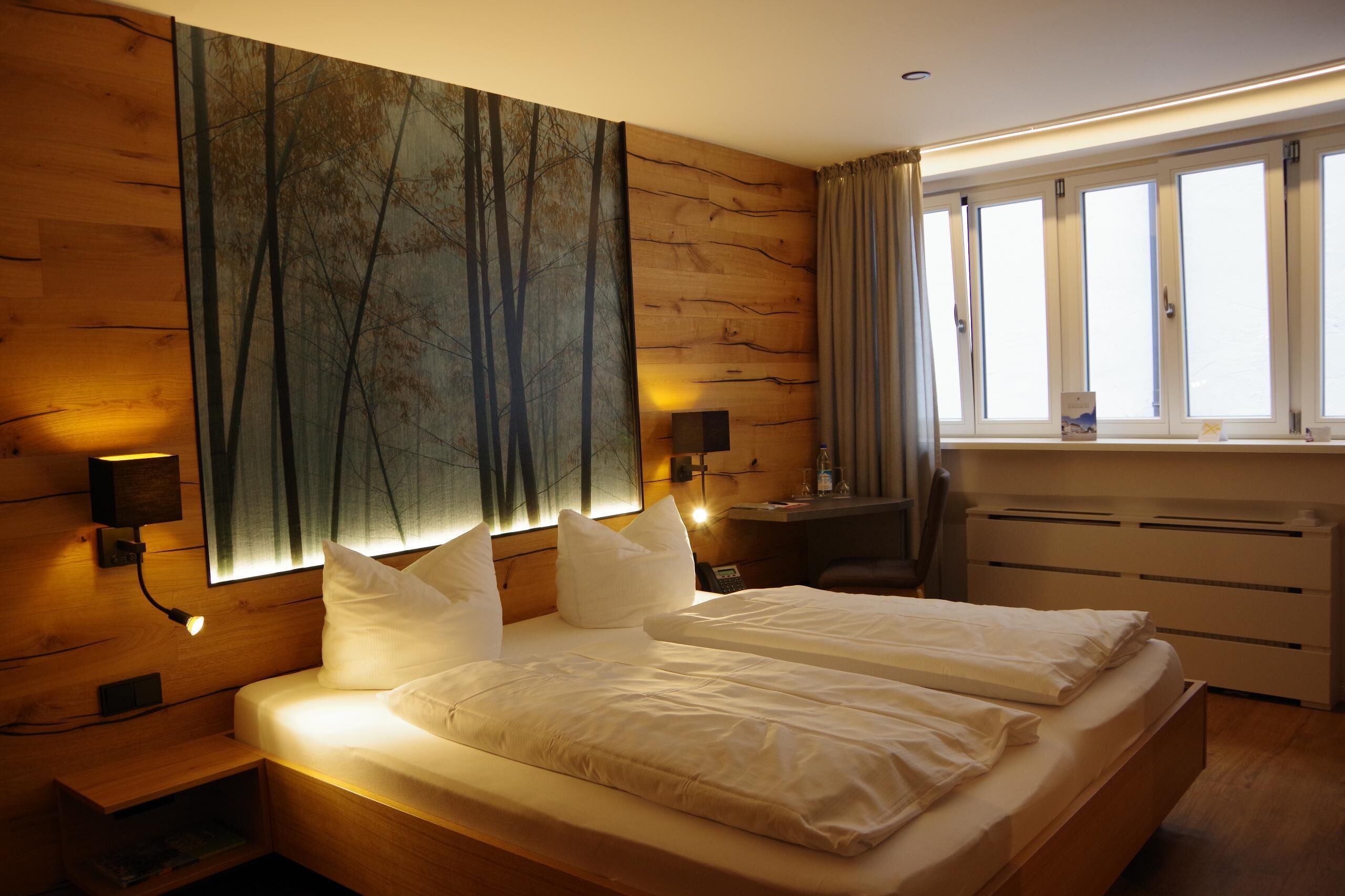 A bedroom with a large bed, a desk, a chair, a nightstand, and a window with curtains.