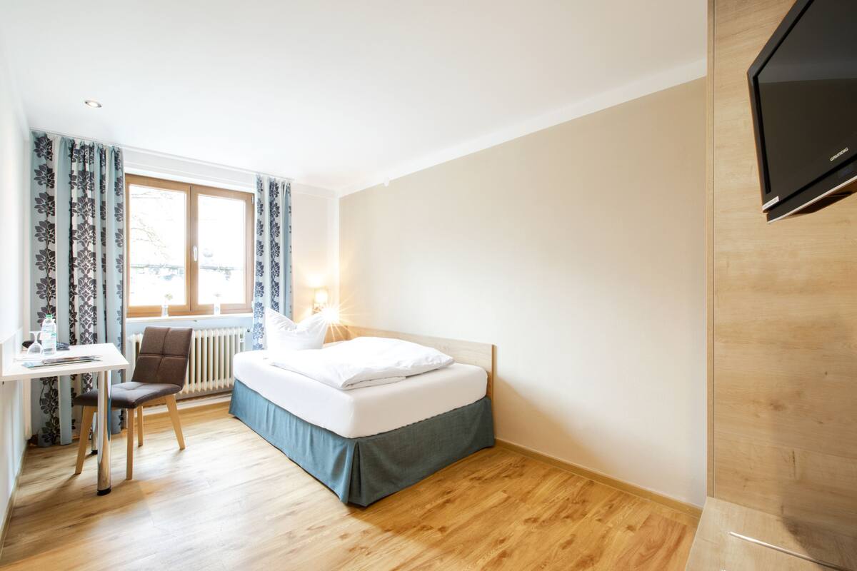A hotel room with a bed, a desk, a chair, a radiator, and a TV.