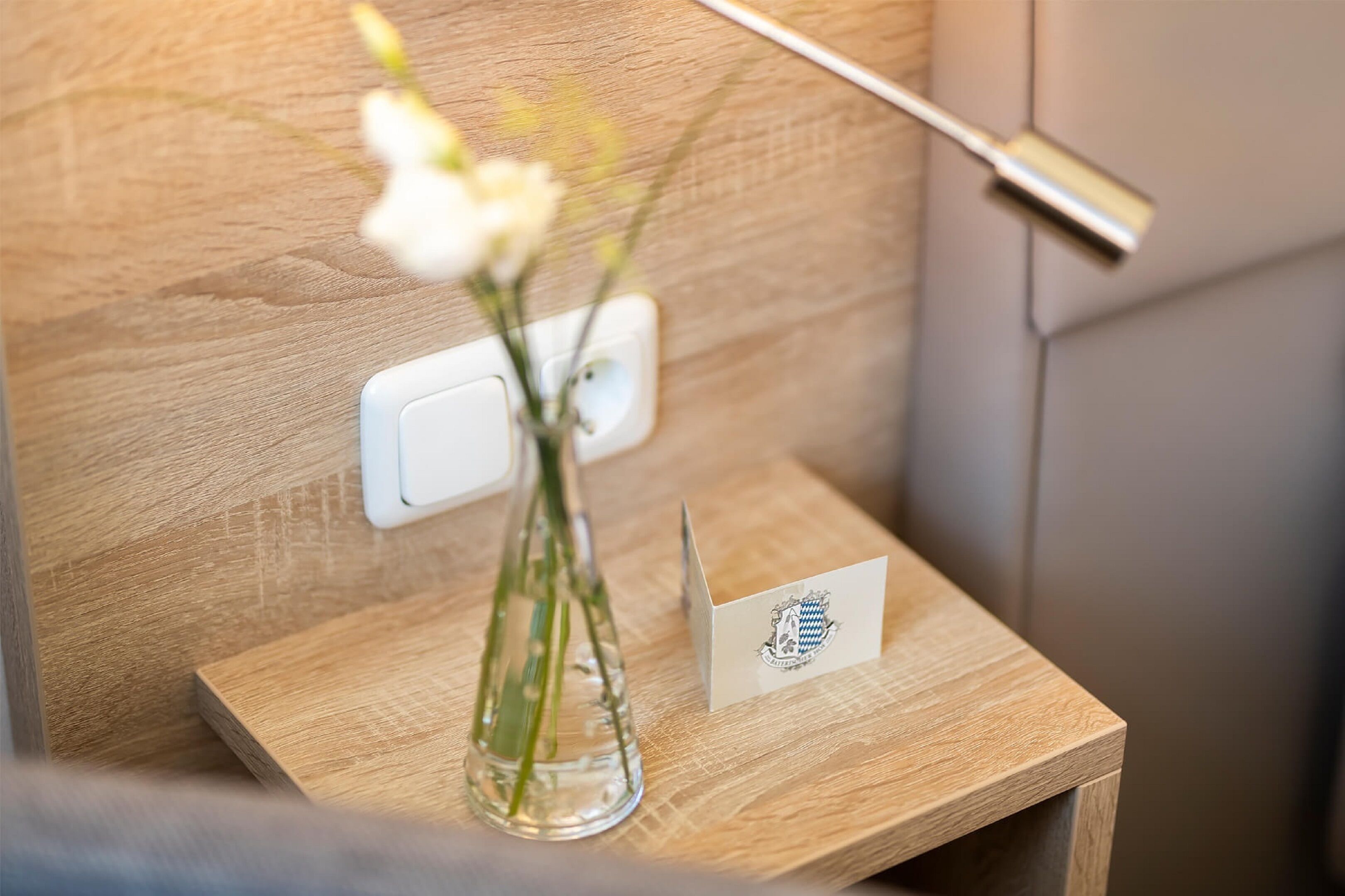 A wooden bedside table with a clear vase containing white flowers, a small card, and a lamp.