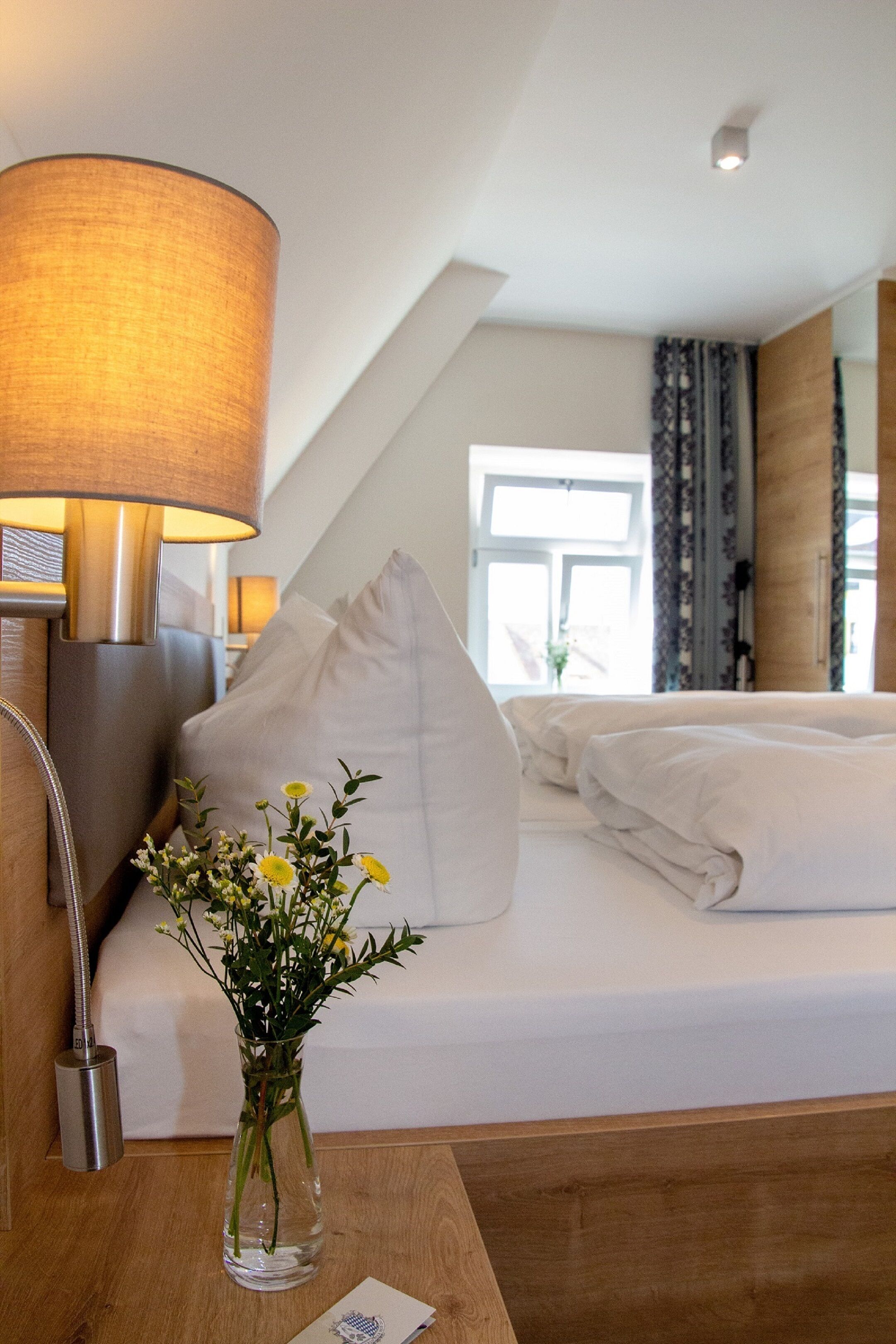 Una habitación de hotel con una cama, una mesita de noche, una lámpara y un florero con flores.