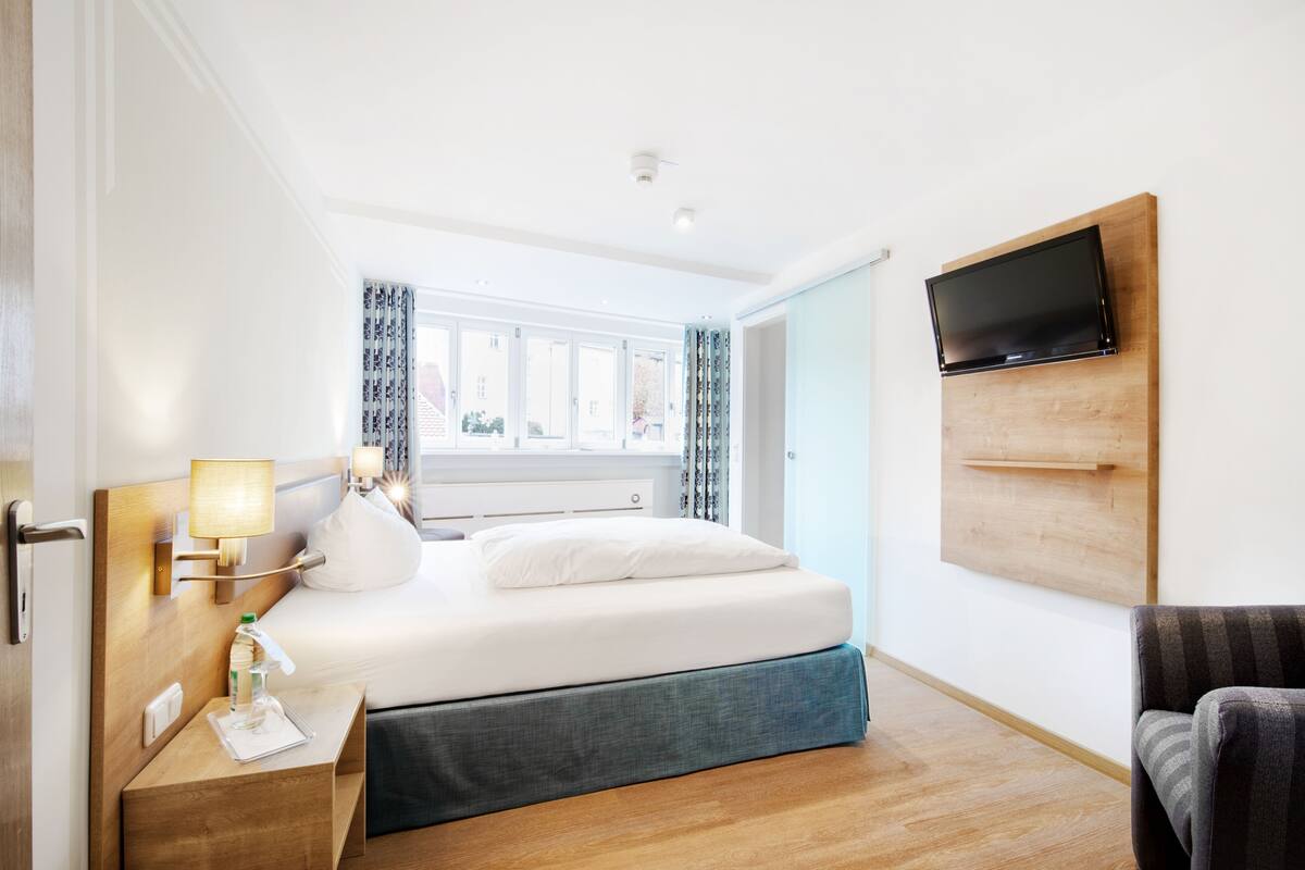 A hotel room with a bed, bedside table, lamp, TV mounted on the wall, and a view of the outside.