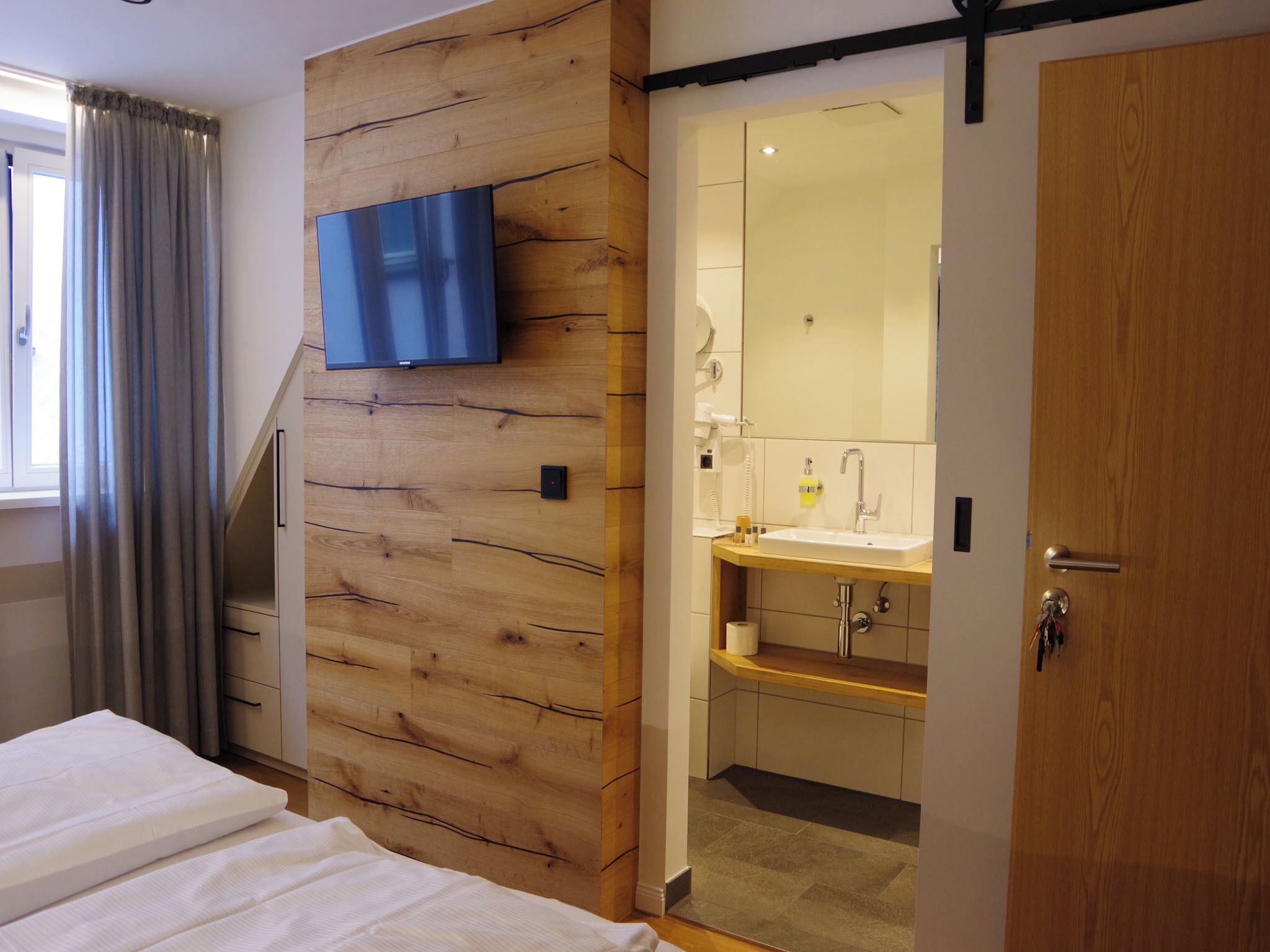 Een slaapkamer met een bed, houten paneelwand, televisie en een badkamer met wastafel en spiegel.