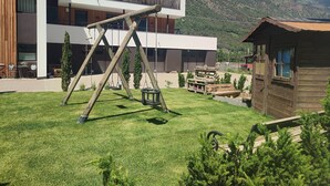 A modern building with a wooden deck, a swing set, and a rustic cabin in a mountainous area.