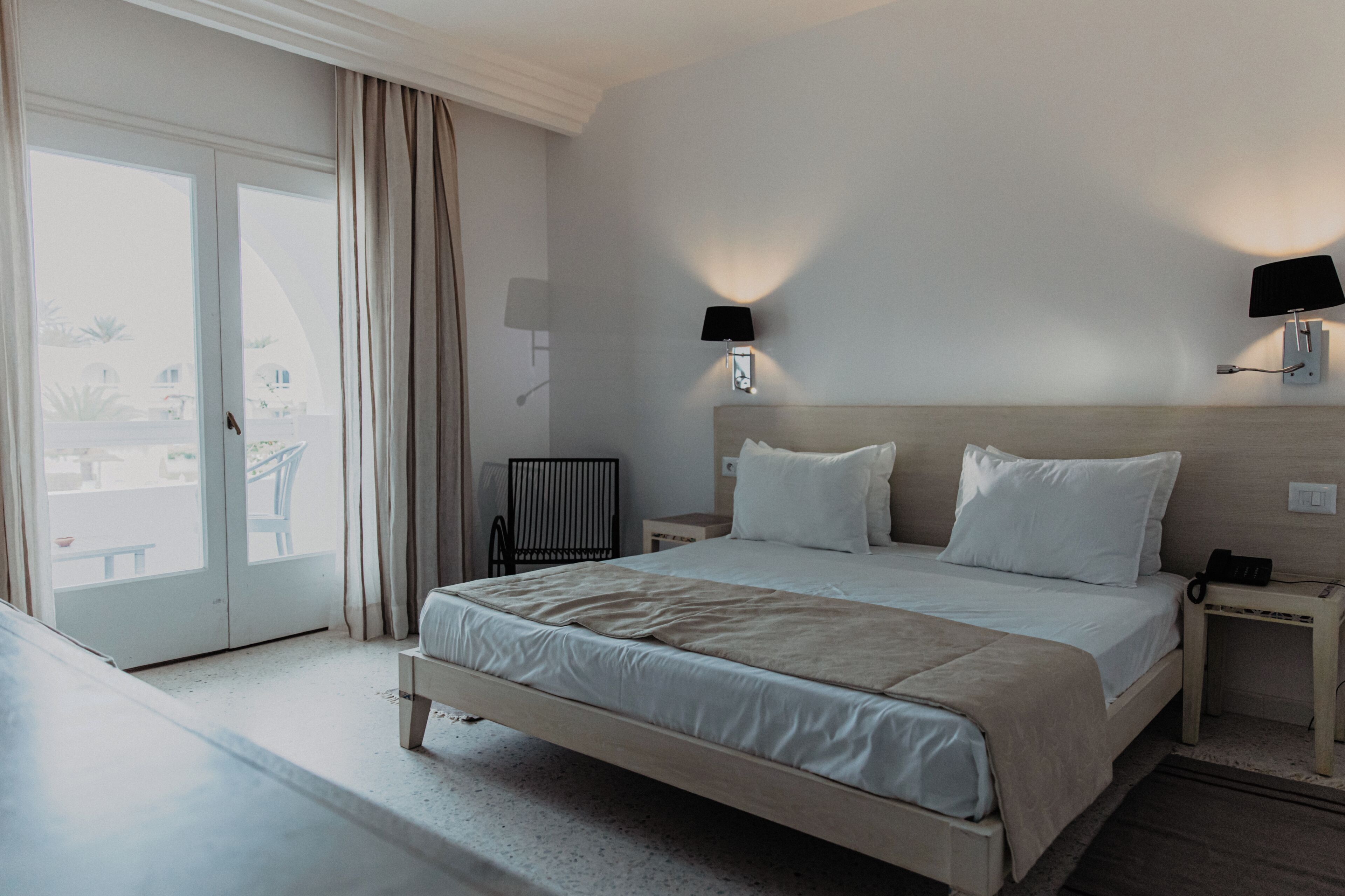Une chambre d’hôtel avec un lit, une table de chevet, une lampe et une vue sur l’extérieur, visible à travers une porte.