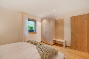 A bedroom with a bed, a wooden wardrobe, a bench, and a window with a view of greenery.