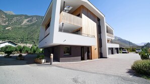 Um edifício moderno de dois andares com varanda, entrada por rampa e montanhas ao fundo.