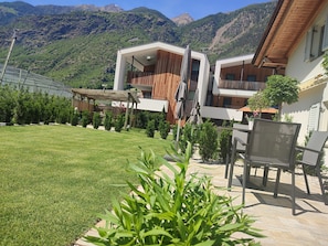 A modern building with a covered outdoor seating area, a well-maintained lawn, and a mountainous backdrop.