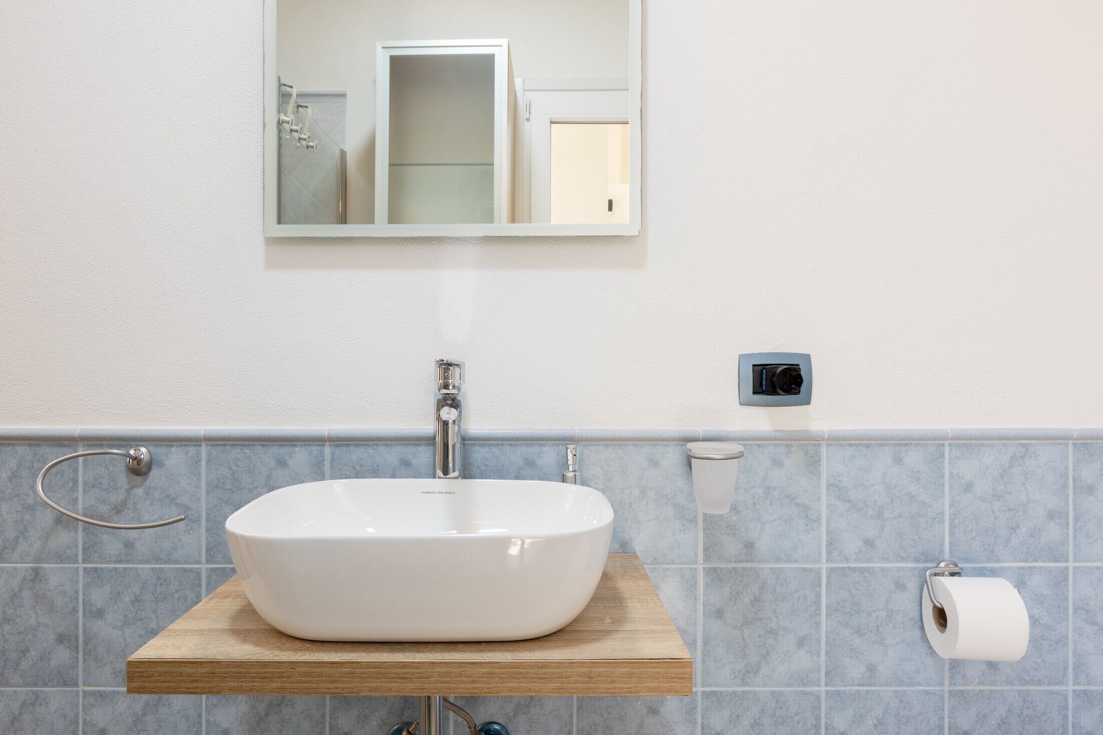 Et badeværelse med en hvid vask, et spejl, et håndklædestativ og en toiletpapirholder.