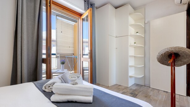 Une chambre d’hôtel avec un lit, un ventilateur sur pied, une fenêtre avec des rideaux et un balcon donnant sur une vue agréable.