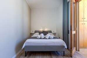 Cama bem arrumada com almofadas e toalhas, mesa de cabeceira com abajur e guarda-roupa de madeira.