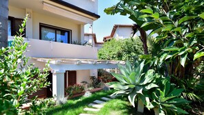 A residential building with a white exterior, a balcony, and a garden with various plants. - Holiday Home "Loft Pina" with Wi-Fi, Garden & Terrace (Arbatax)