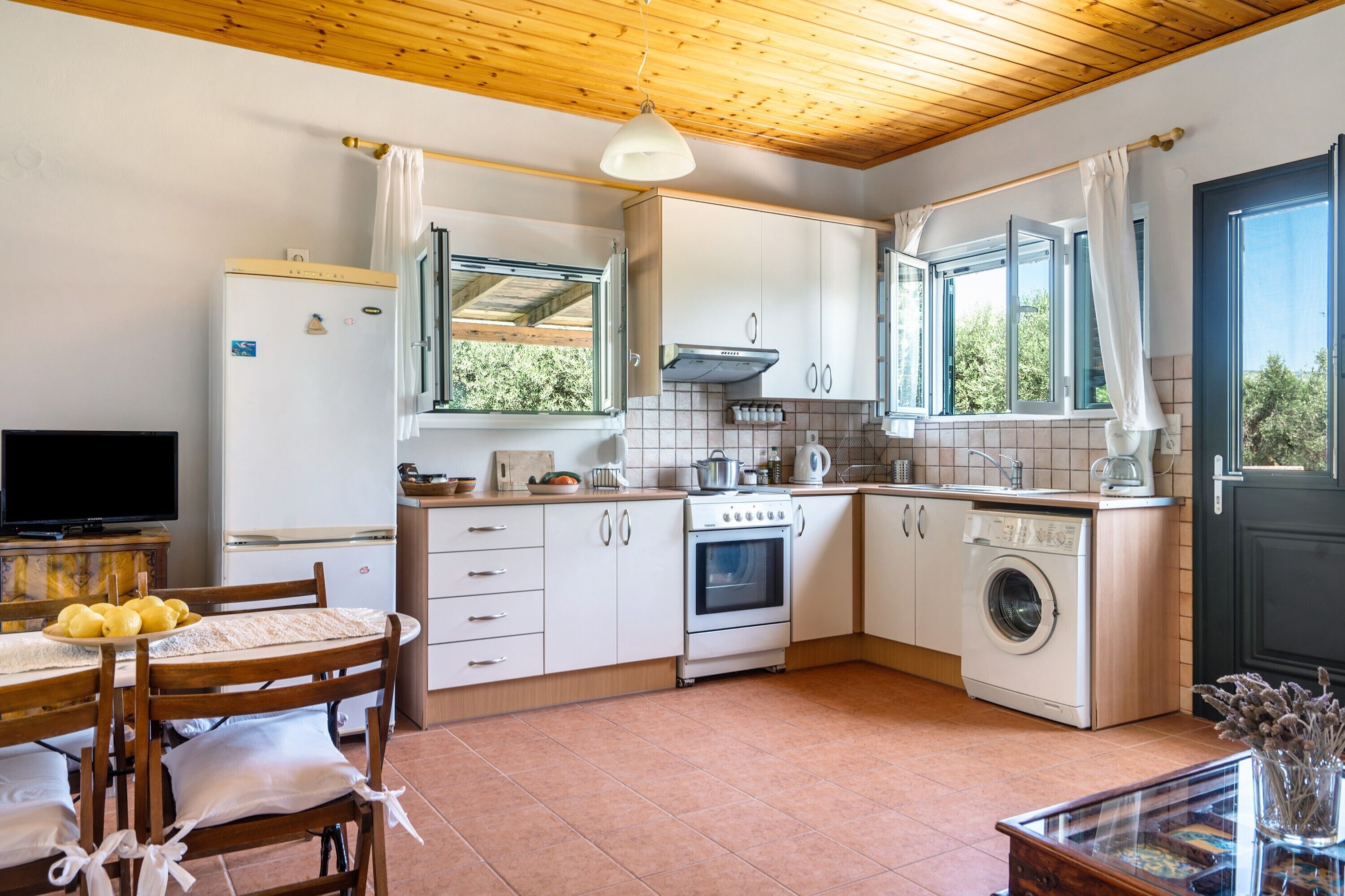Une cuisine compacte avec un plafond en bois, des armoires blanches, un four encastré, un lave-linge et un coin repas avec une table et des chaises.