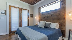 Un dormitorio con cabecera de madera, una cama grande con colcha azul y blanca, una ventana con persianas y una mesita de noche con lámpara.