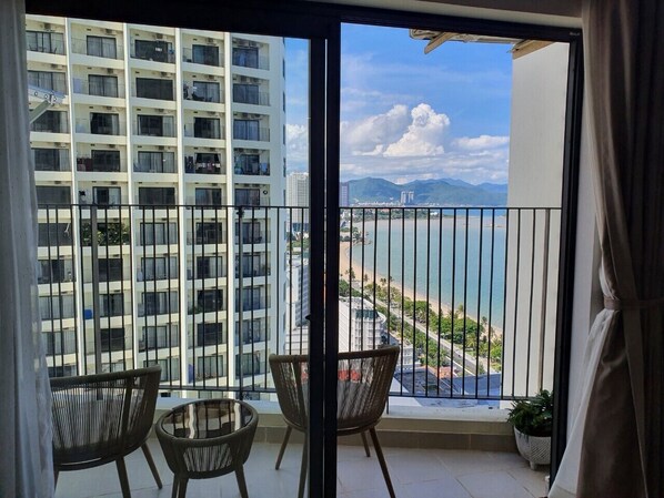 A balcony with two wicker chairs and a small table, overlooking a beach and a cityscape with high-rise buildings.