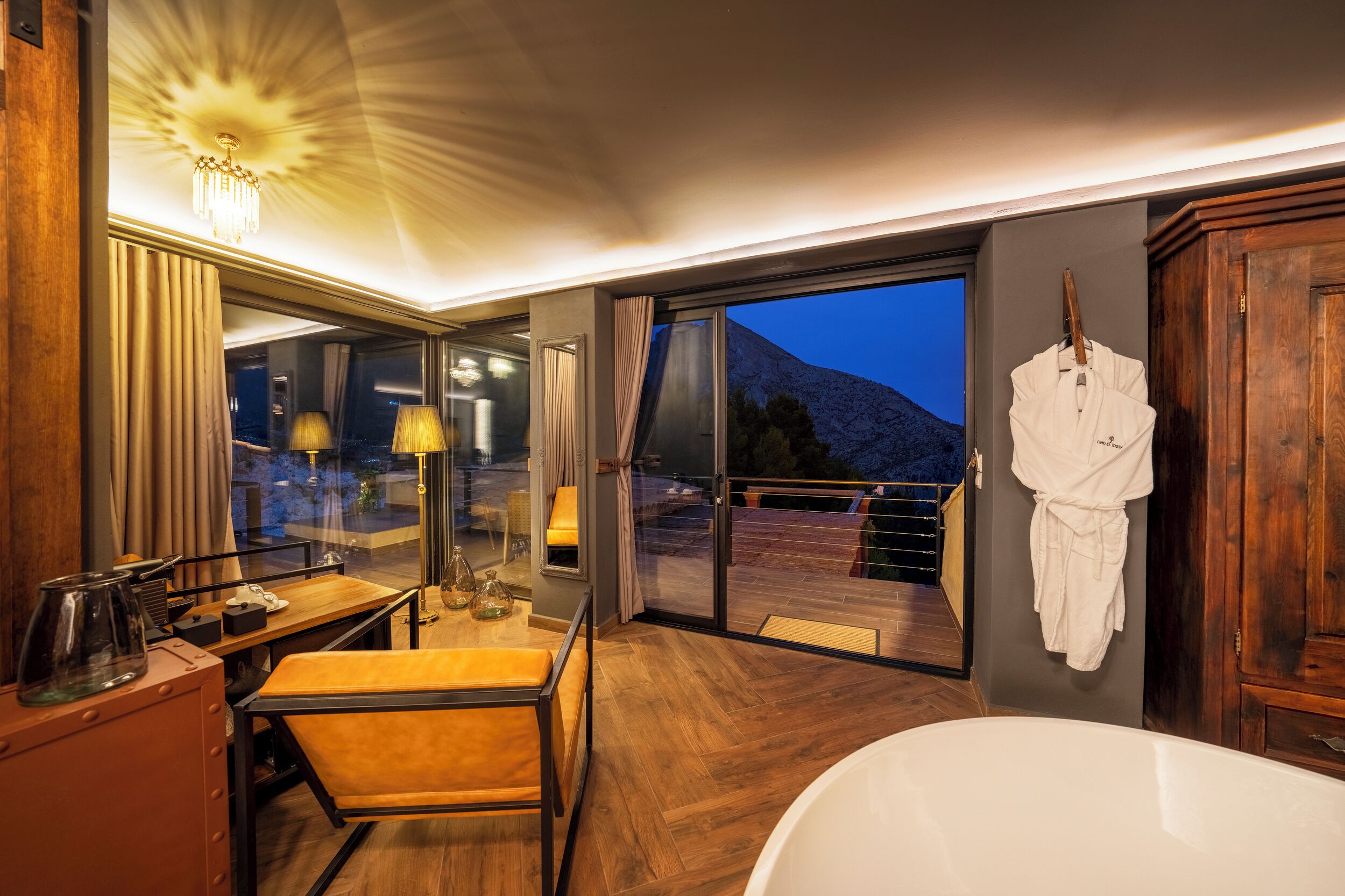 Un baño moderno con una bañera grande, suelo de madera, un espejo y vista a una montaña al atardecer.
