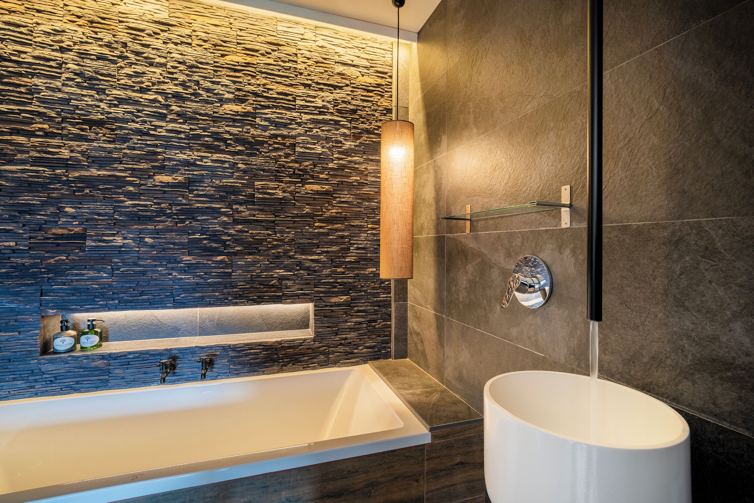Un baño moderno con pared de piedra, una bañera blanca, un estante de vidrio con artículos de aseo y un lavabo redondo.