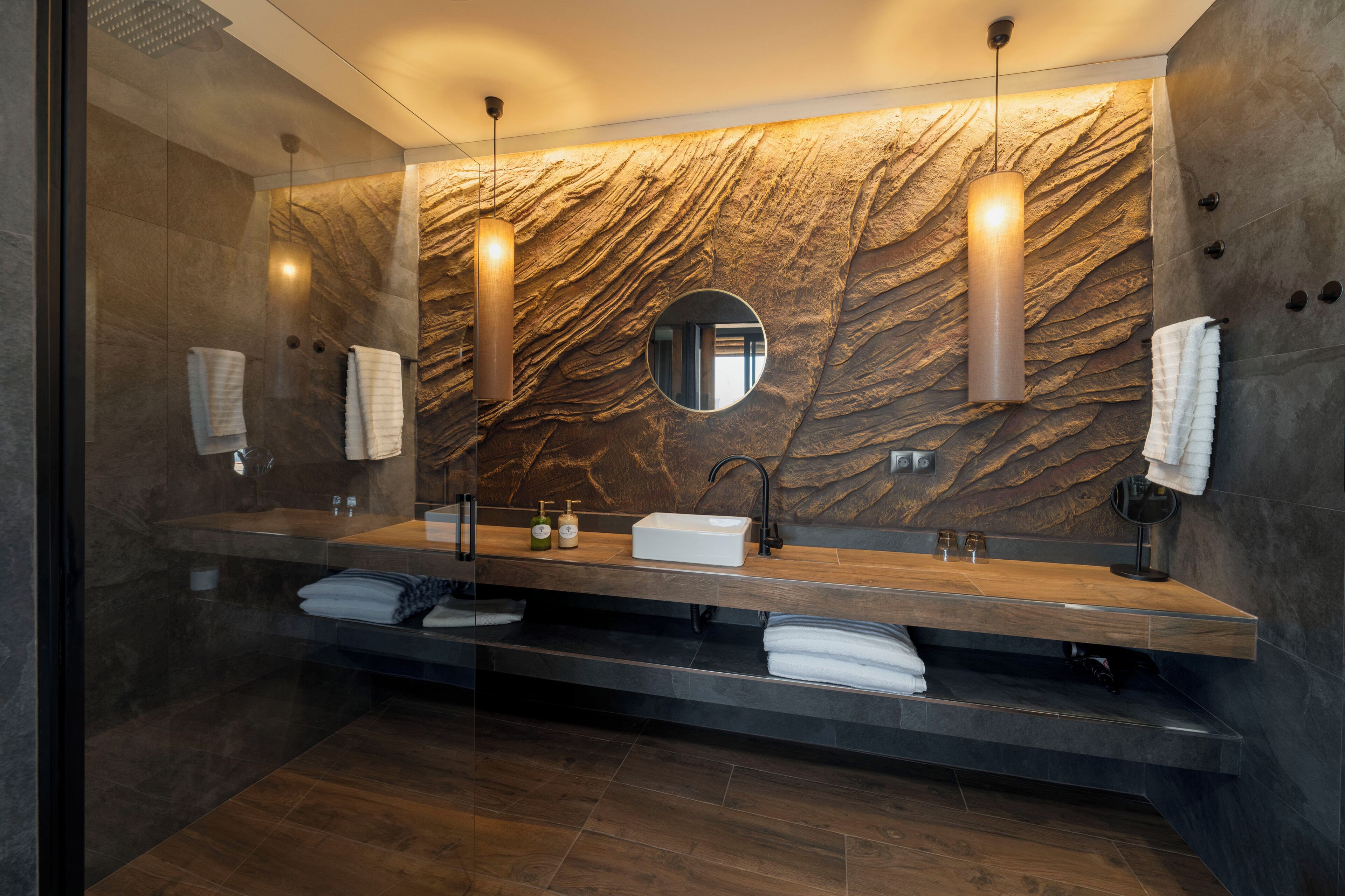 A modern bathroom with a marble wall, a large mirror, and a wooden vanity.