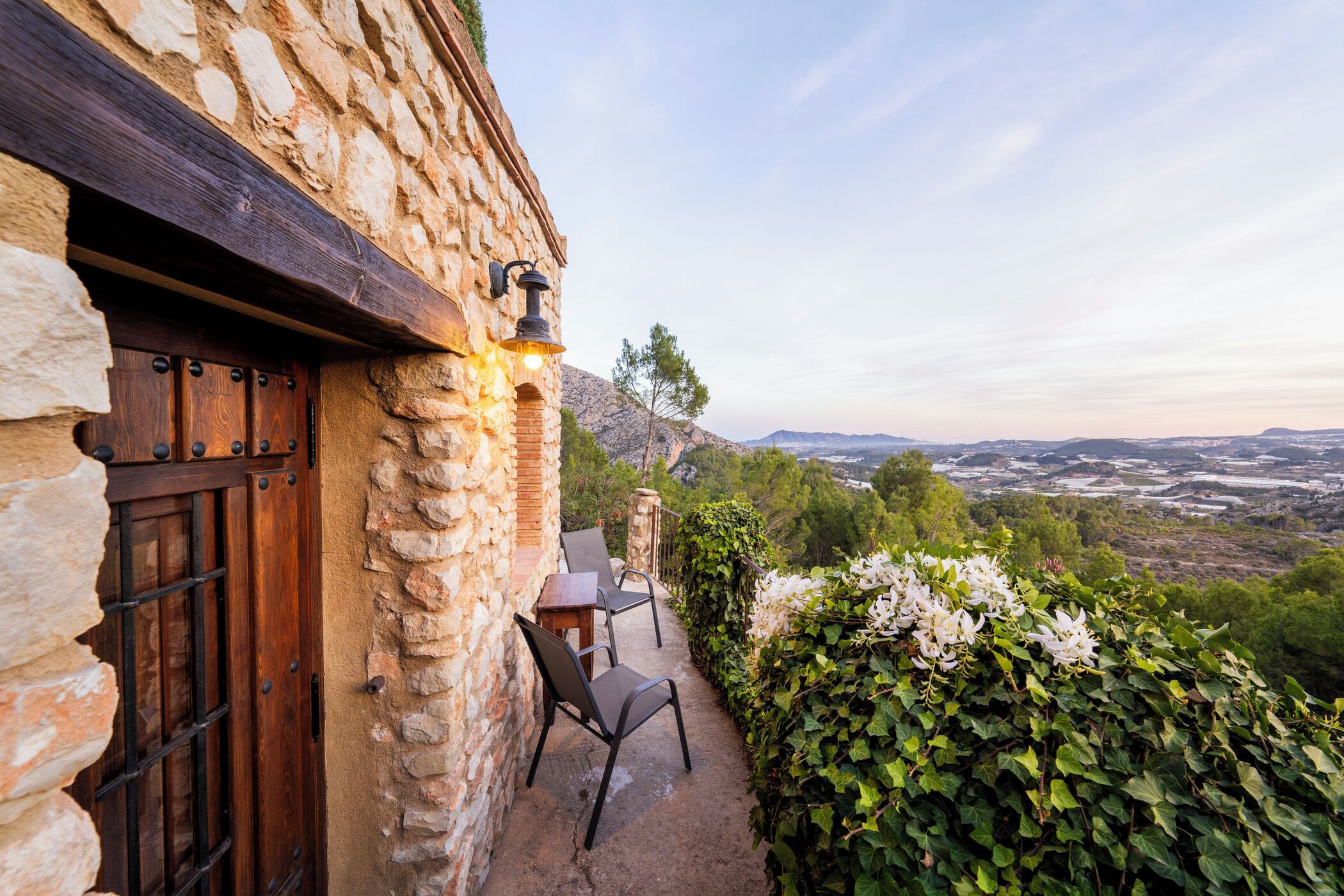 A stone building with a wooden door, outdoor seating, and a view of a valley.