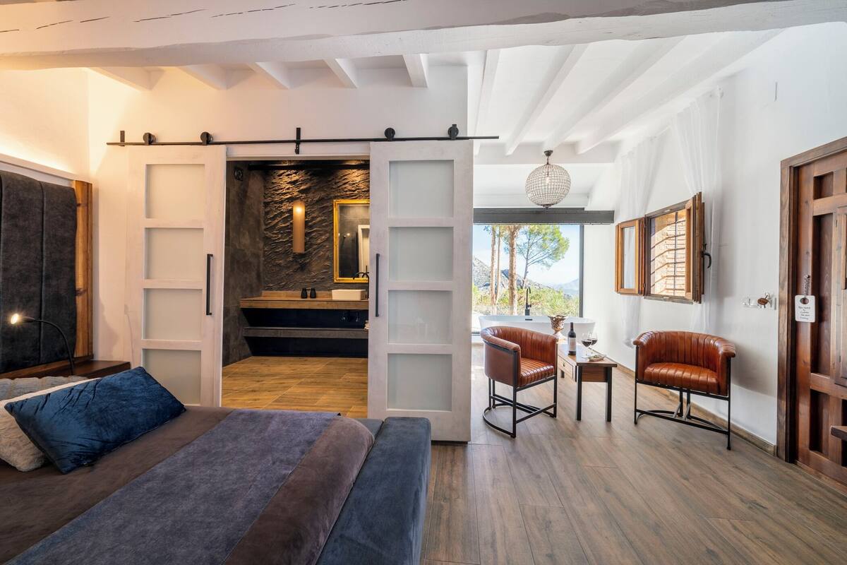 Un dormitorio moderno con cama, escritorio, silla y vistas a un muro de piedra y paisaje exterior.