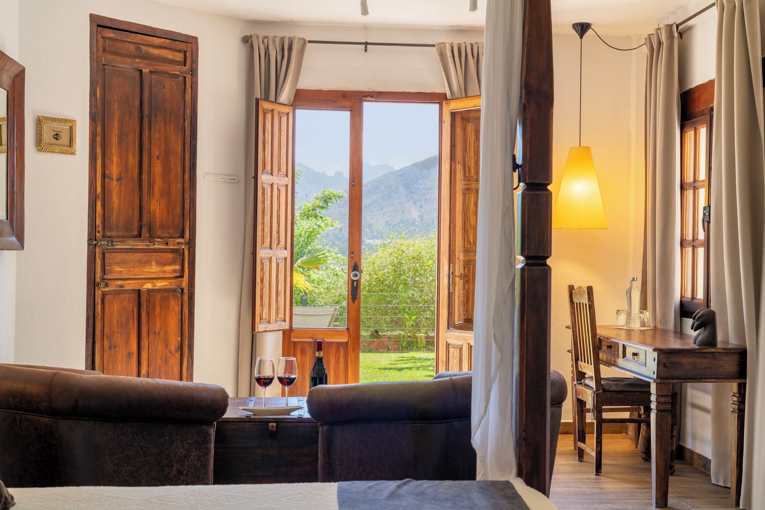Una stanza con un letto, un tavolo di legno con due bicchieri di vino rosso, una sedia e vista sulle montagne attraverso porte aperte.