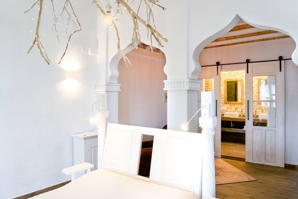 Un dormitorio con cama, pared blanca, suelo de madera y un arco blanco que da a un baño.