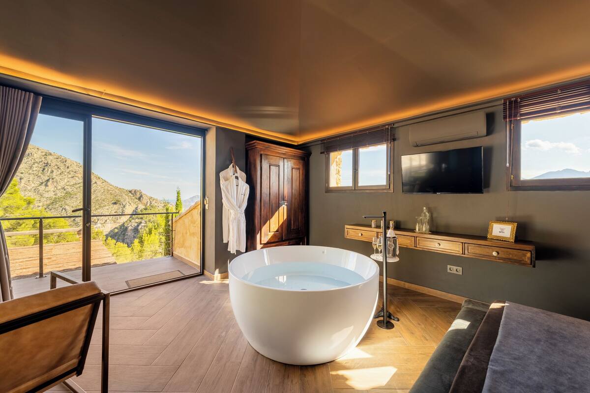 Un baño moderno con una bañera independiente grande, un tocador de madera, un televisor montado y un balcón con vistas a las montañas.