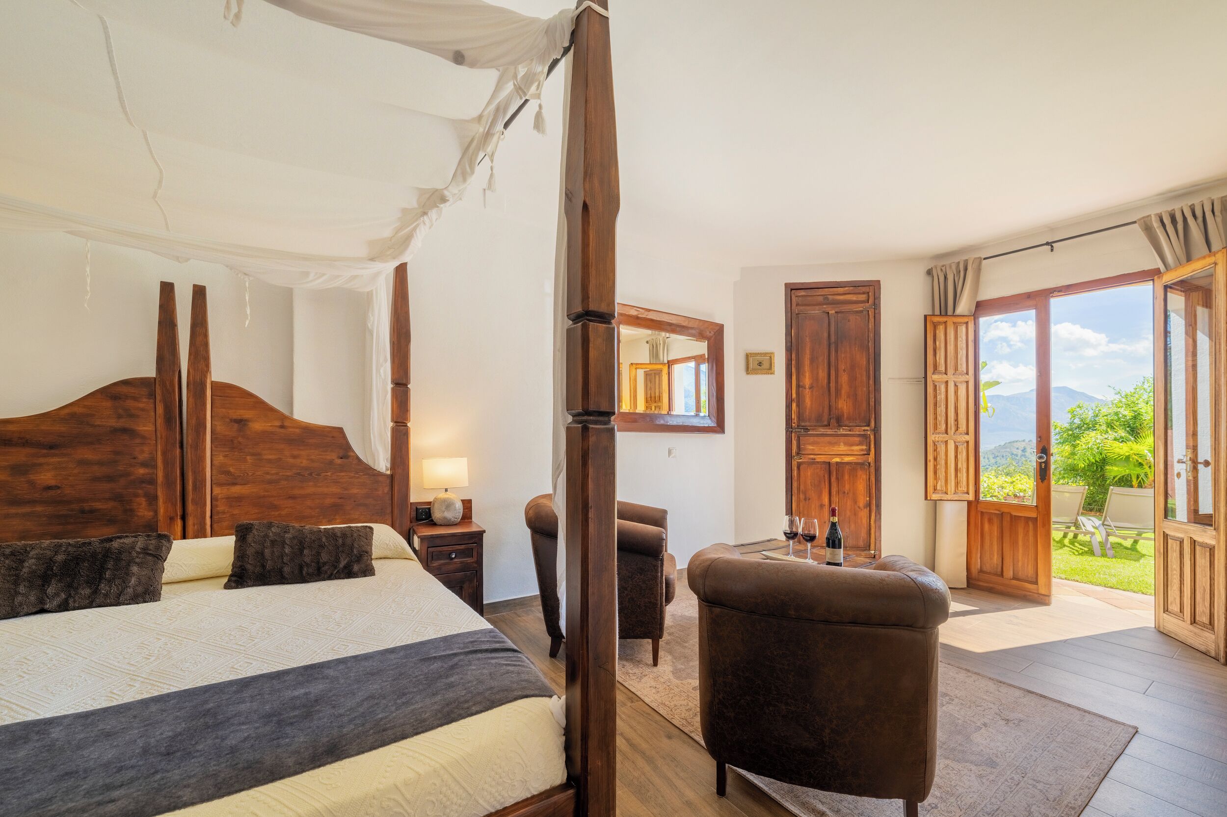 A bedroom with a large bed, a wooden headboard, a nightstand, a chair, and a view of the outdoors through a large window.
