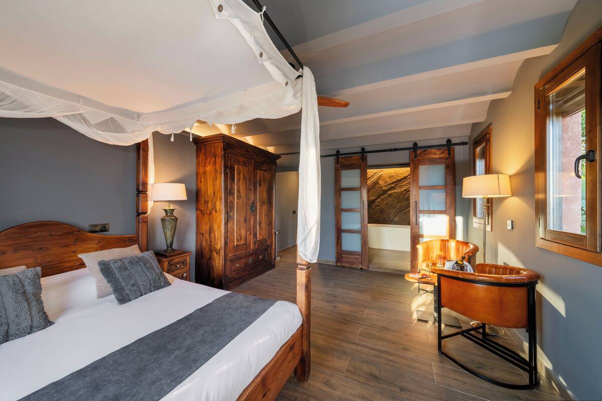 Un dormitorio con cama con dosel, armario de madera y una zona de estar con una silla y una mesa.