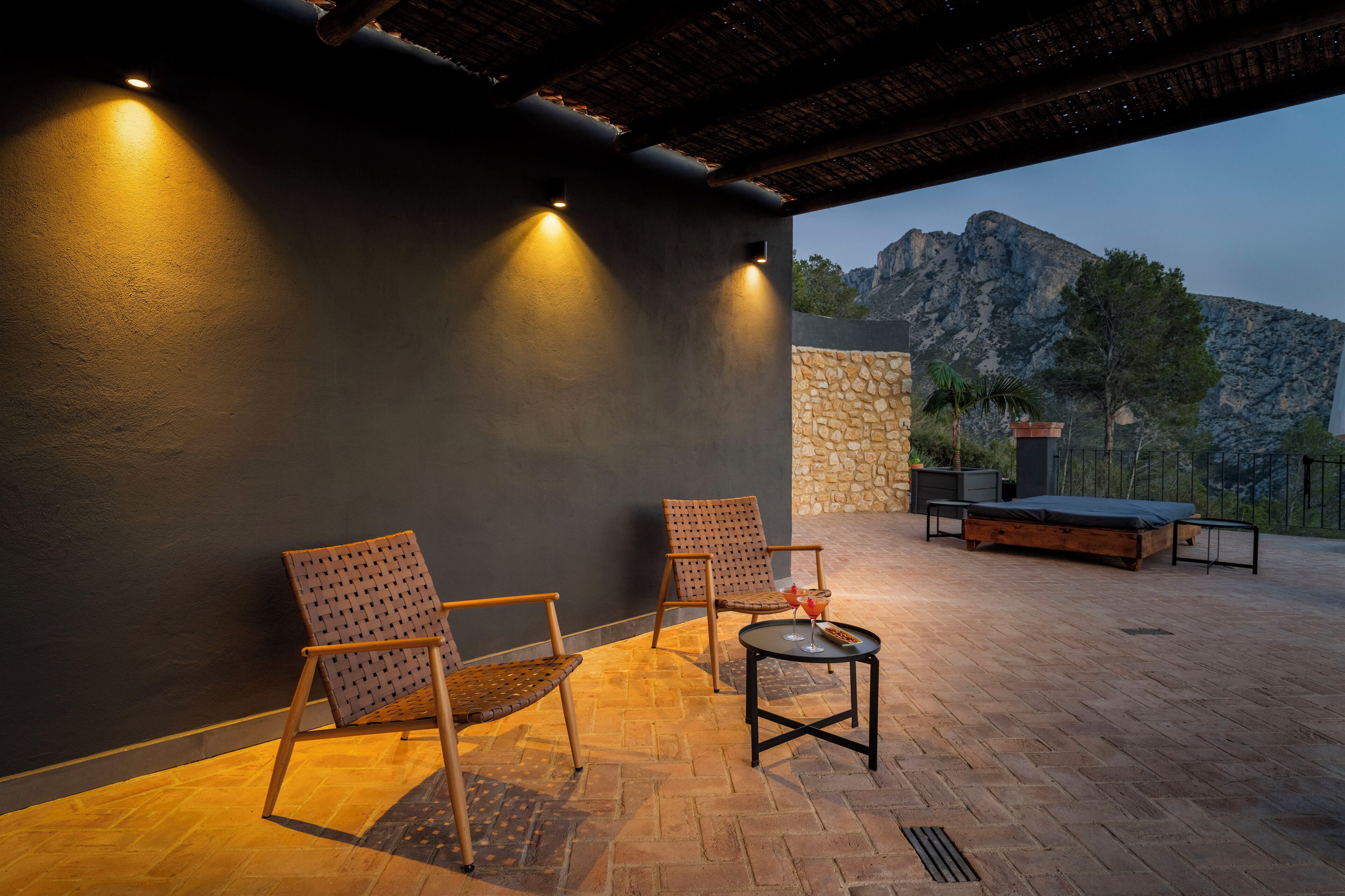 Un patio al aire libre con dos sillas de mimbre, una mesita redonda con una bebida y una cama.