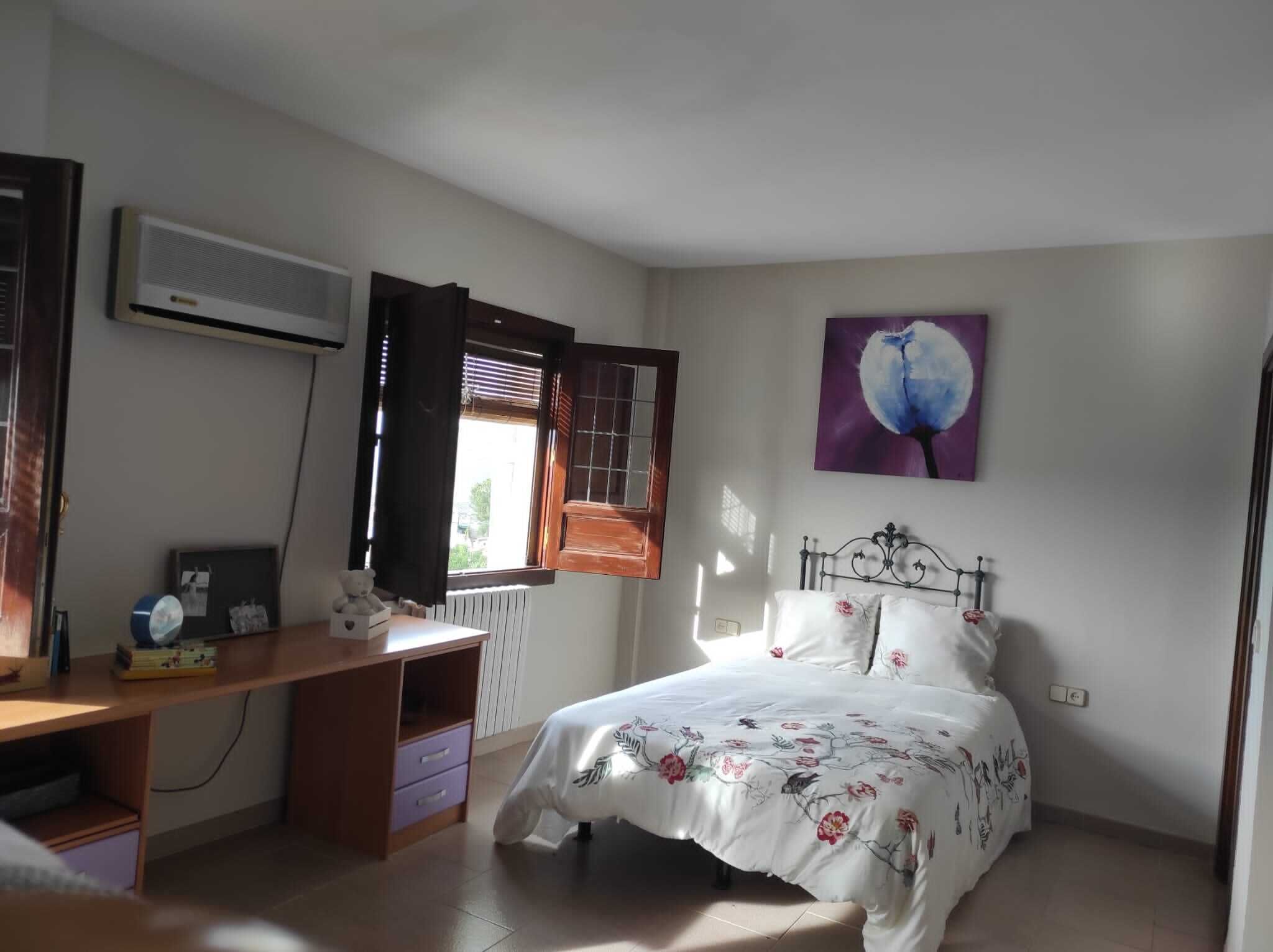 Ein Schlafzimmer mit Bett, Schreibtisch, Heizkörper, einem Fenster mit Vorhängen und einem Bild an der Wand.
