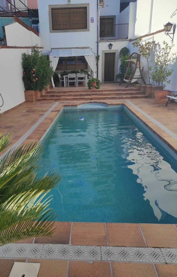 A rectangular swimming pool with a tiled deck, surrounded by a white building with a balcony and a small outdoor seating area.