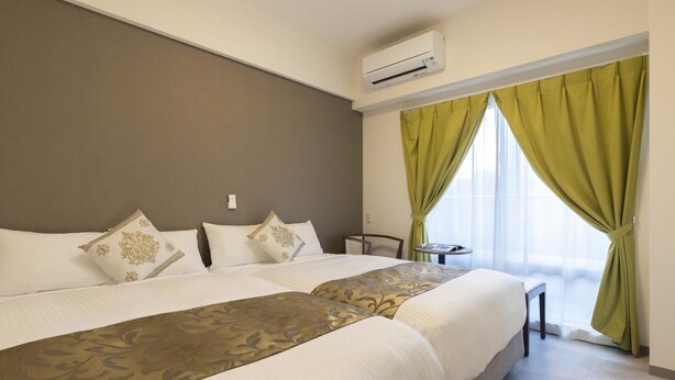 Ein Hotelzimmer mit zwei Betten, einem kleinen Tisch, einem Stuhl und einem Fenster mit grünen Vorhängen.