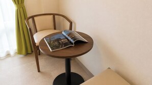 A small round table with a book, a chair, and a bench in a room with a plain wall and a window with curtains. - Mr.KINJO GRAND CATS (Ishigaki)