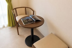 A small round table with a book, a chair, and a bench in a room with a plain wall and a window with curtains. - Mr.KINJO GRAND CATS (Ishigaki)