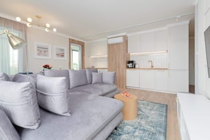 A modern living room with a grey sectional sofa, a round wooden coffee table, and a kitchen area with white cabinets and a wooden countertop.