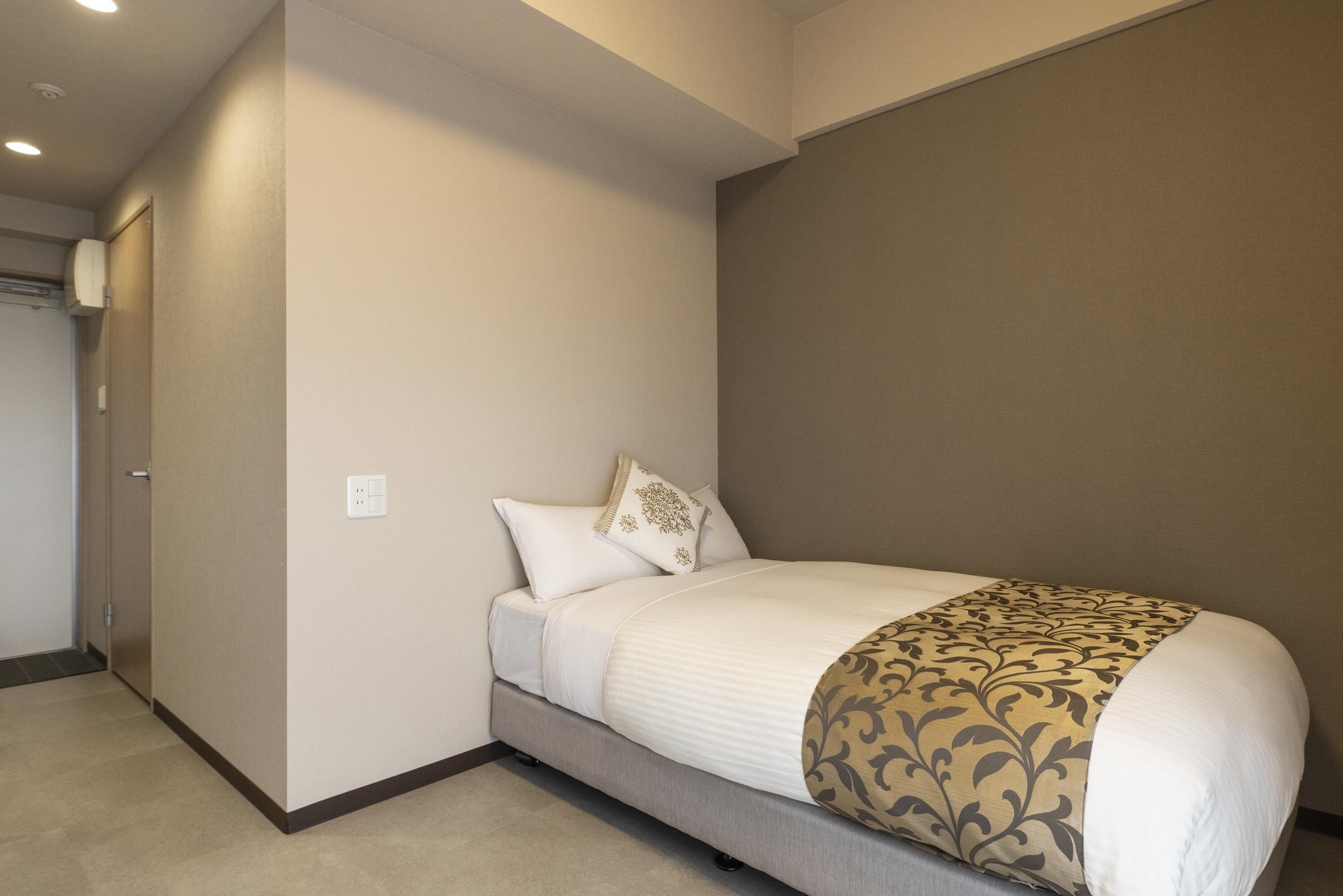 A neatly made bed with a patterned throw, a plain wall, and a door to the left.