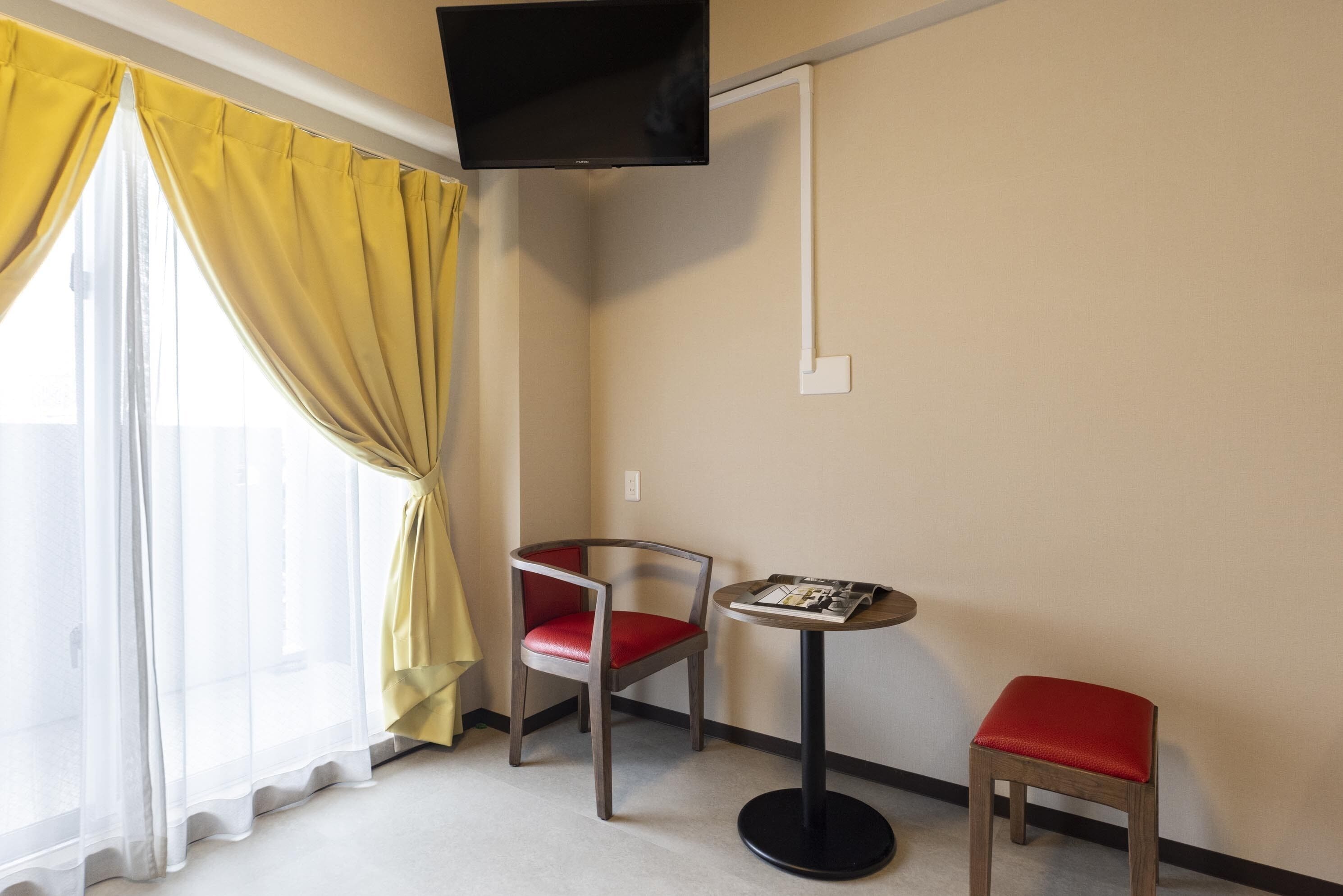 A hotel room with a single bed, a TV mounted on the wall, a small table with a magazine, a chair with a red cushion, and a stool with a red cushion.